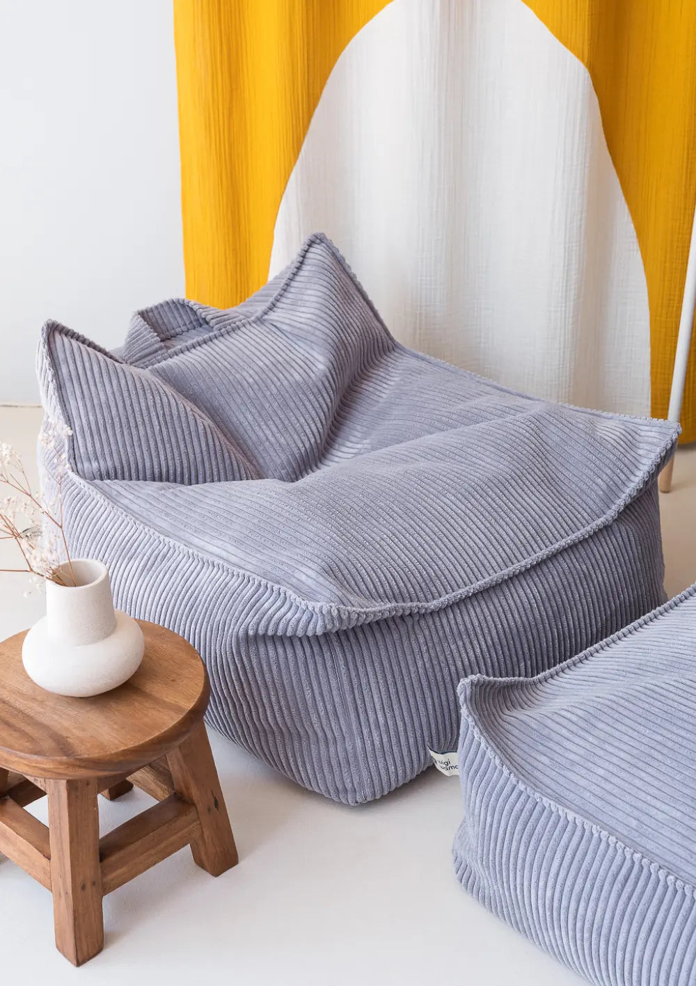 Der taubenblaue Wigiwama Sitzsack Cord Blueberry Blue aus weichem Breitcord steht freistehend neben einem kleinen Holzschemel in einem hellen Kinderzimmer.