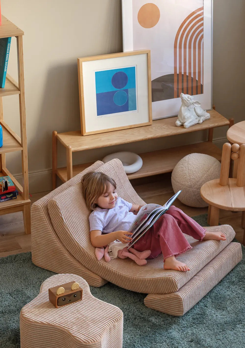 Ein Kind liegt zurückgelehnt im hellbraunen Wigiwama Kindersessel Moon Chair Cord Brown Sugar aus Breitcord und liest aufmerksam ein Bilderbuch.
