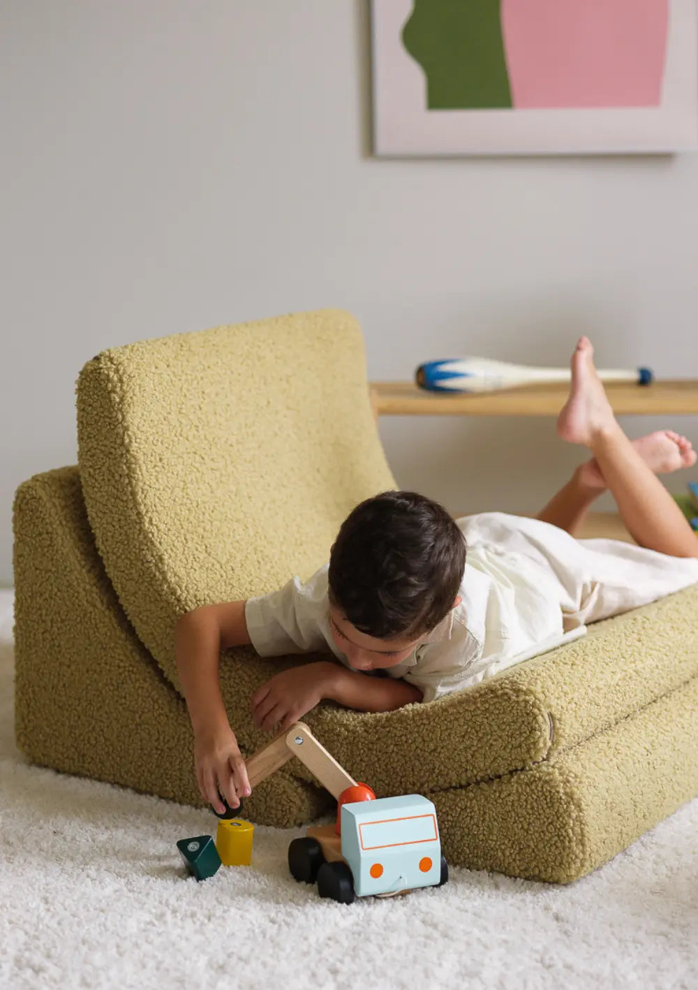 Ein Kind liegt Bbuchlings auf dem aufgeklappten Wigiwama Kindersessel Moon Chair Teddy Matcha und spielt mit einem Holzspielzeug.