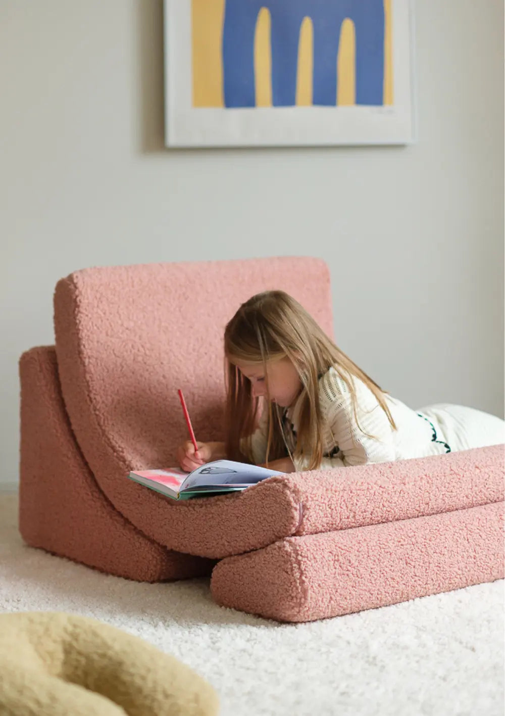 Ein Mädchen liegt bauchlings im aufgeklappten, altrosafarbener Wigiwama Kindersessel Moon Chair Teddy Guava und malt mit einem Stift in ein Buch.