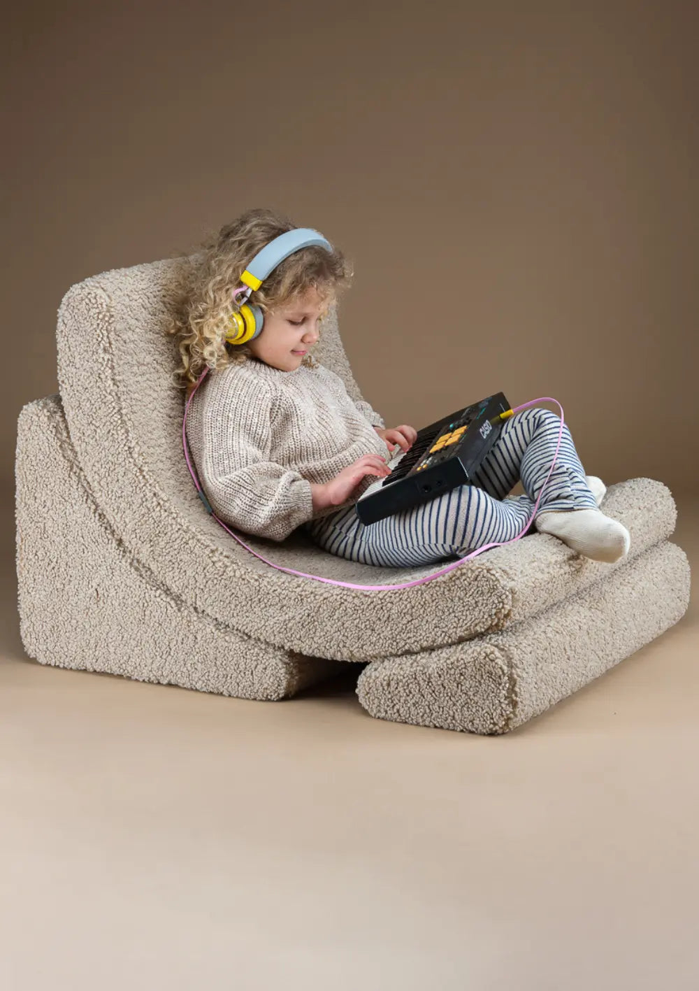 Ein Kind mit gelben Kopfhörern sitzt gemütlich im beigefarbenen Wigiwama Kindersessel Moon Chair Teddy Biscuit aus Teddy-Plüsch und spielt auf einem Mini-Keyboard.