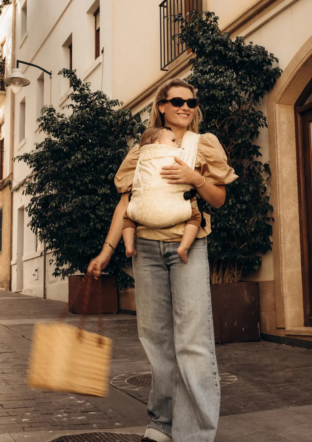 Eine lächelnde Frau mit Sonnenbrille und Korbtasche blickt beim Gehen auf einer Straße zur Seite, während ihr Baby sanft vor ihrer Brust in der beigefarbenen Wildride Babytrage Firstride Bowie mit Leopardmuster ruht.