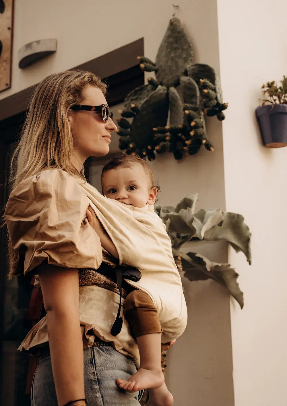 Seitenansicht einer Frau, die ihr Baby vor der Brust in der hellen Wildride Babytrage Firstride Bowie Beige Leopard mit dezentem Tiermuster trägt, während das Kind neugierig in die Kamera blickt.