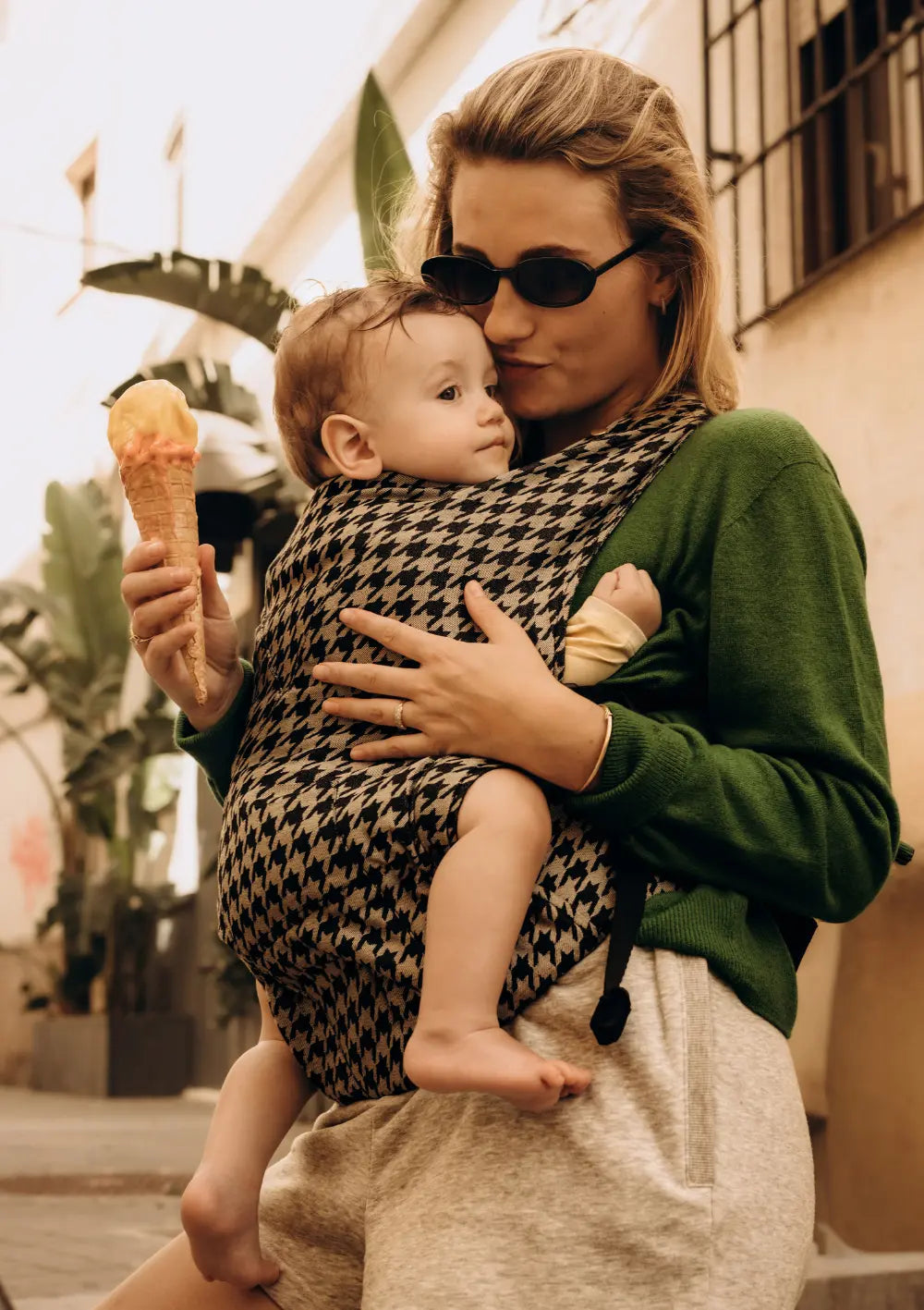 Eine Frau mit Sonnenbrille und grüne Pullover hält ein Waffeleis in der Hand und trägt ihr Baby vor der Brust in der Wildride Babytrage Firstride Bowie mit klassischem Hahnentritt-Muster.
