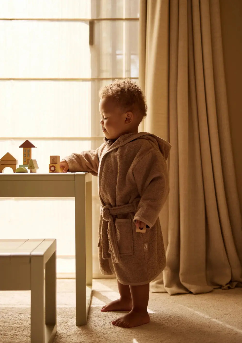 Ein Kind im braunen Bademantel steht am Kindertisch und spielt mit den Tieren und Bausteinen des Jollein Holz Bauklötze Jollies Sets.