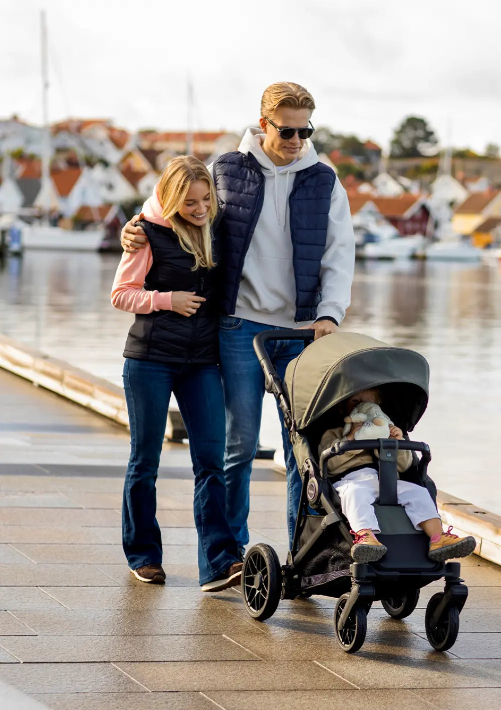 Ein Paar spaziert Arm in Arm an einer Promenade und schiebt den olivgrünen Emmaljunga Kinderwagen Sonic Stroller Urban Green mit sitzendem Kleinkind.