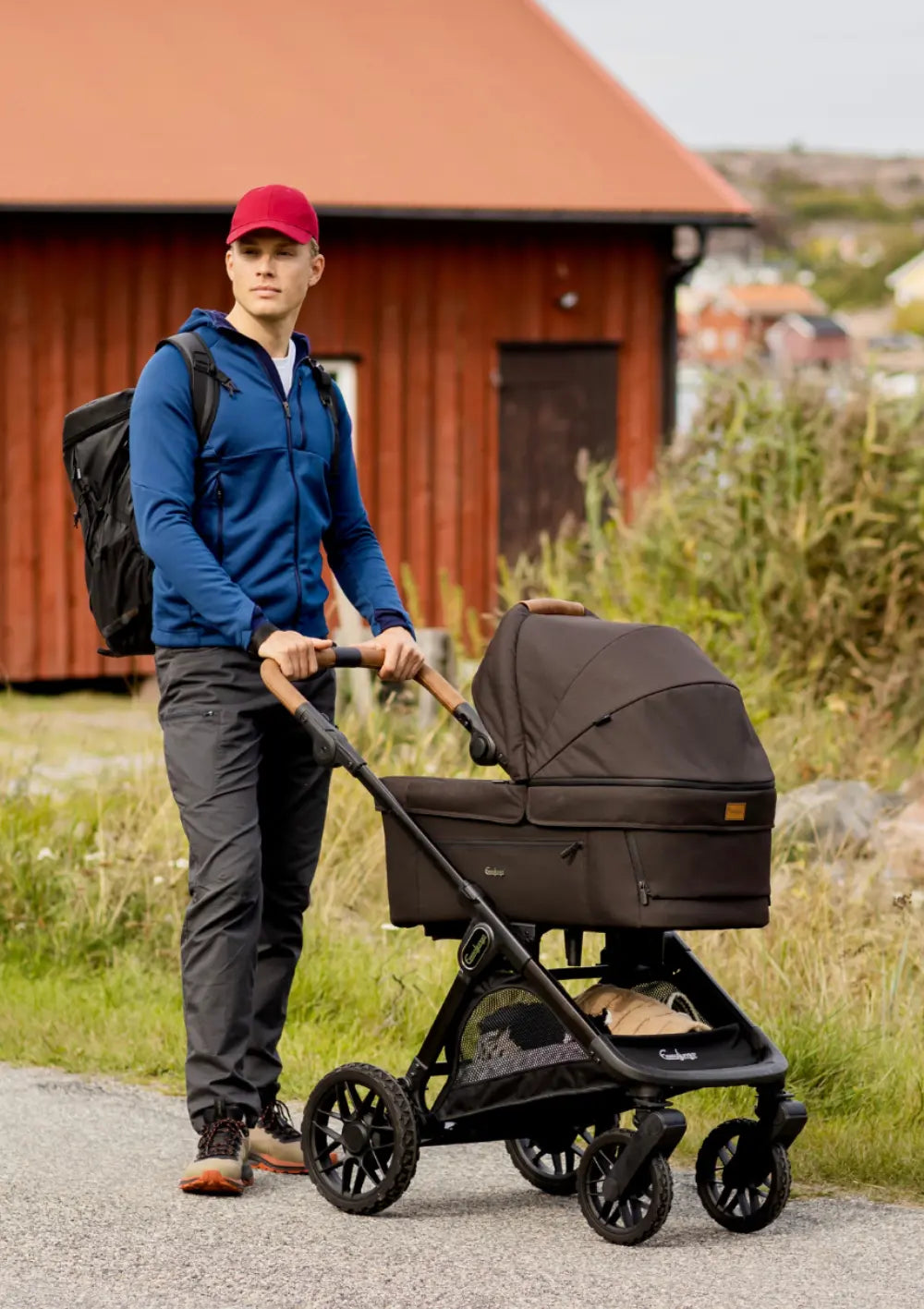 Ein Mann mit roter Cappy und Rucksack schiebt den dunkelbraunen Emmaljunga Kinderwagen SENTO Outdoor Brown auf einem Weg im Freien vor einem roten Holzhaus.