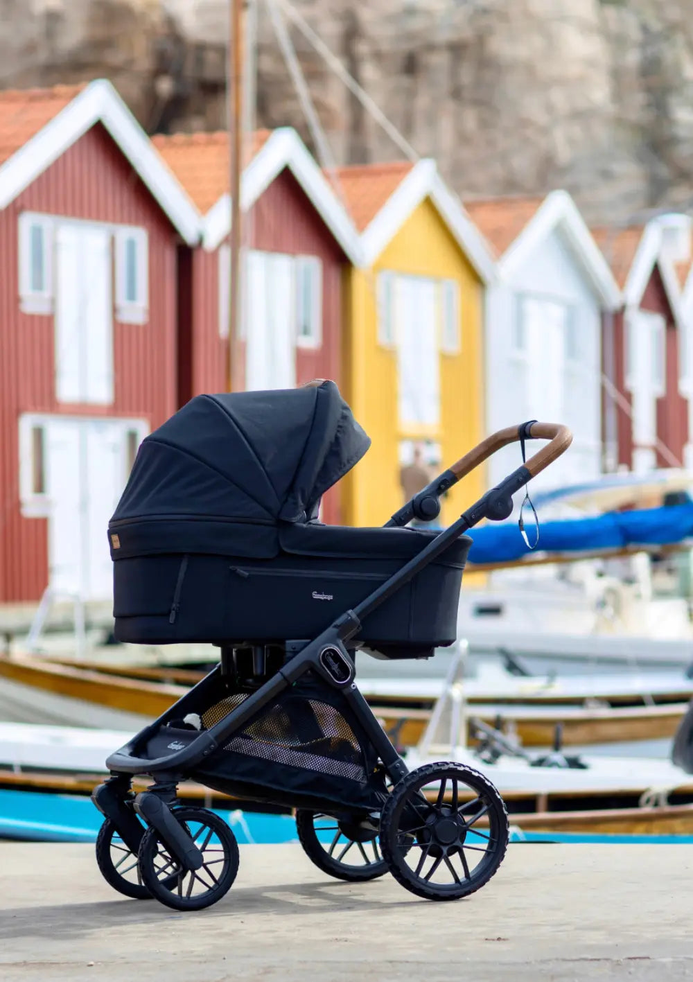 Der dunkelblaue Kombikinderwagen mit Babywanne aus dem Emmaljunga Kinderwagen-Set SENTO PRO Outdoor Navy steht freistehend am Hafen vor bunten Fischerhäusern.