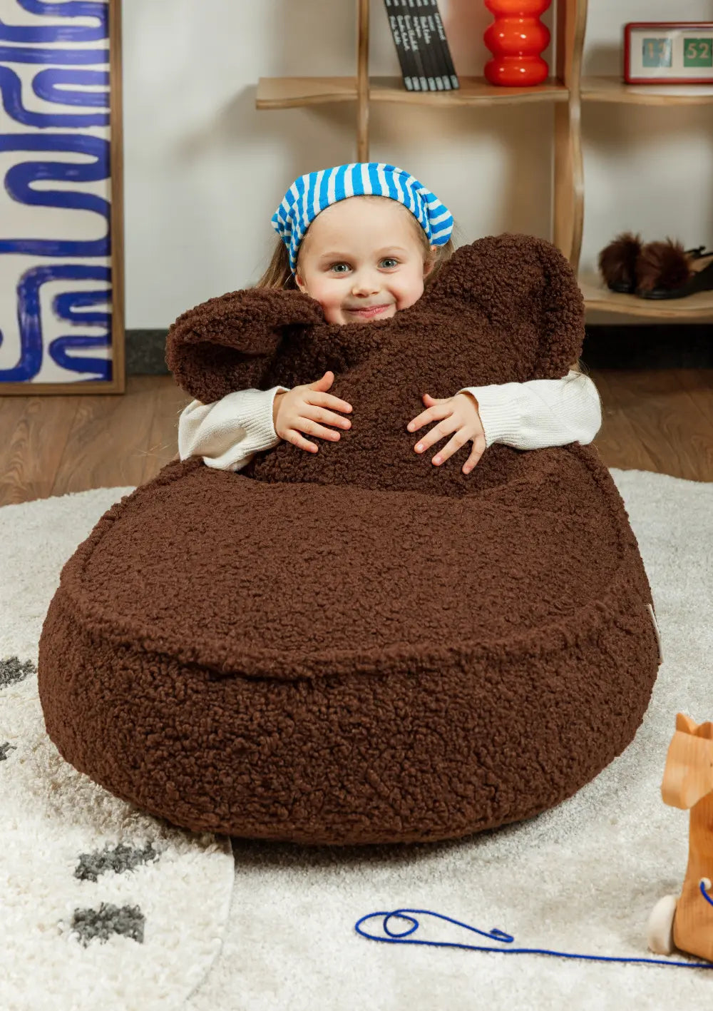 Ein lächelndes Kind umfasst im Kinderzimmer liebevoll die Rückenlehne mit Bärenohren des braunen Wigiwama Bären Sitzsack Teddy Warm Hazel.