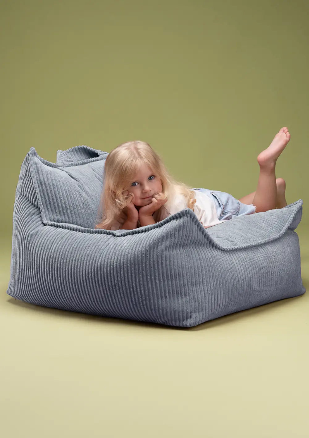 Ein Mädchen liegt bauchlings entspannt im taubenblauen Wigiwama Sitzsack Cord Blueberry Blue aus Breitcord und blickt direkt in die Kamera.