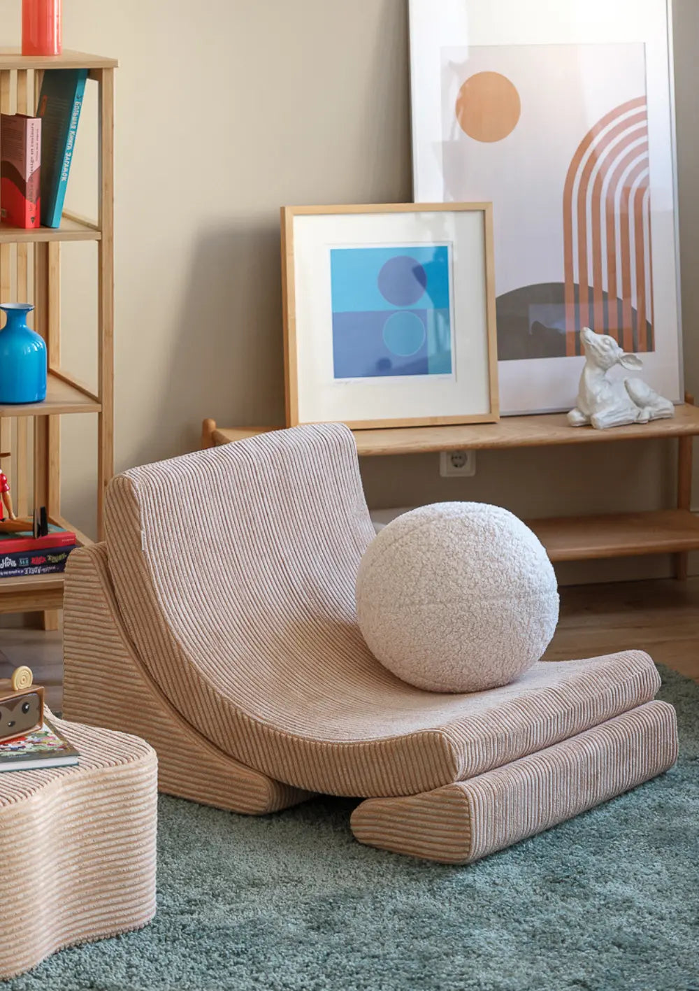 Der hellbraune Wigiwama Kindersessel Moon Chair Cord Brown Sugar aus Breitcord steht zusammen mit einem runden Plüschkissen auf einem grünen Teppich.