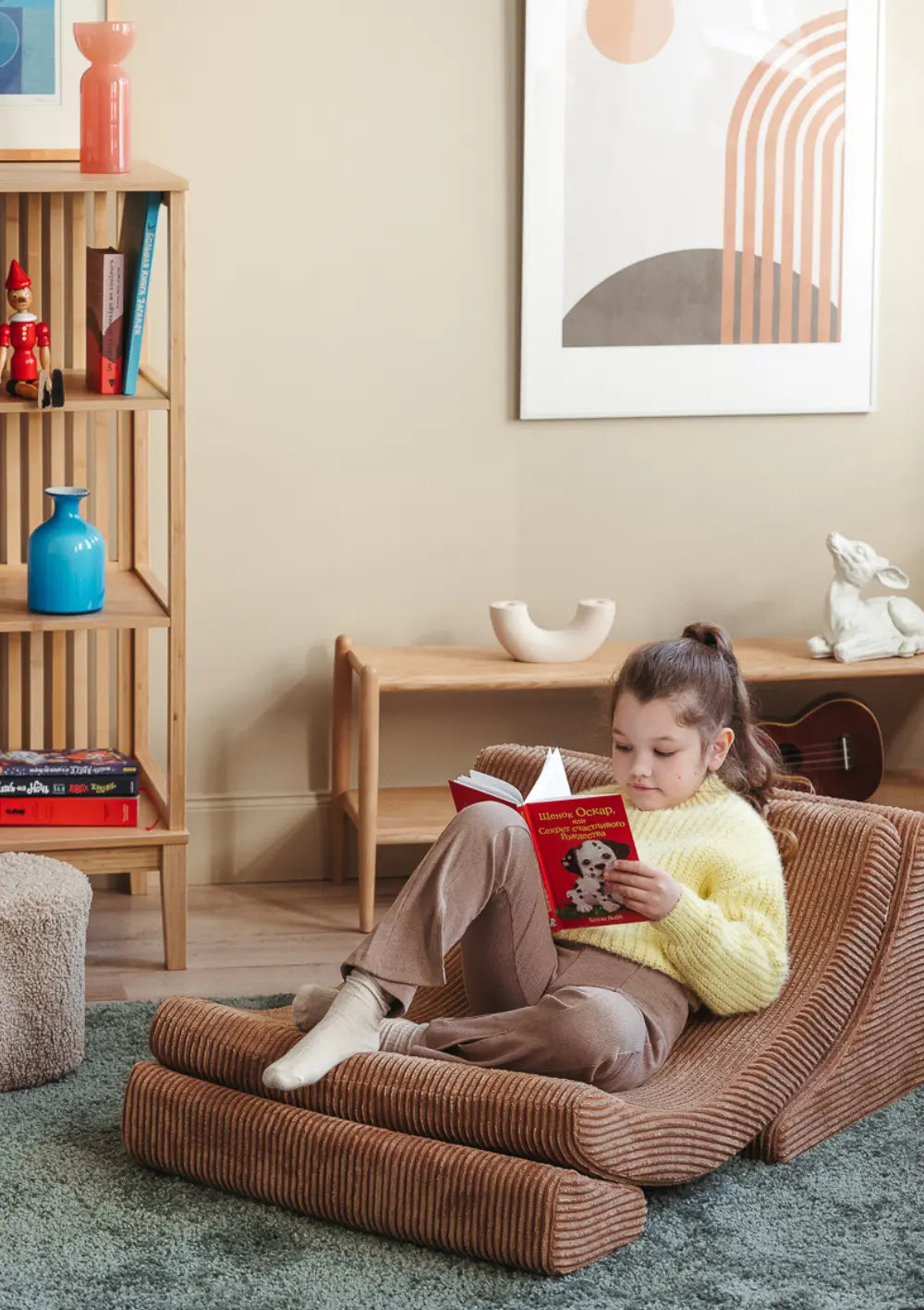Ein Mädchen im gelben Pullover sitzt im braunen Wigiwama Kindersessel Moon Chair Cord Toffee und liest ein Buch.