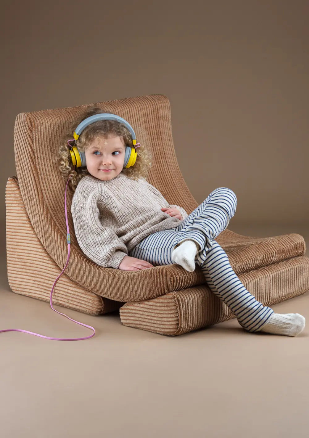 Ein Kind mit gelben Kopfhörern entspannt zurückgelehnt im Wigiwama Kindersessel Moon Chair Cord Toffee aus Breitcord.