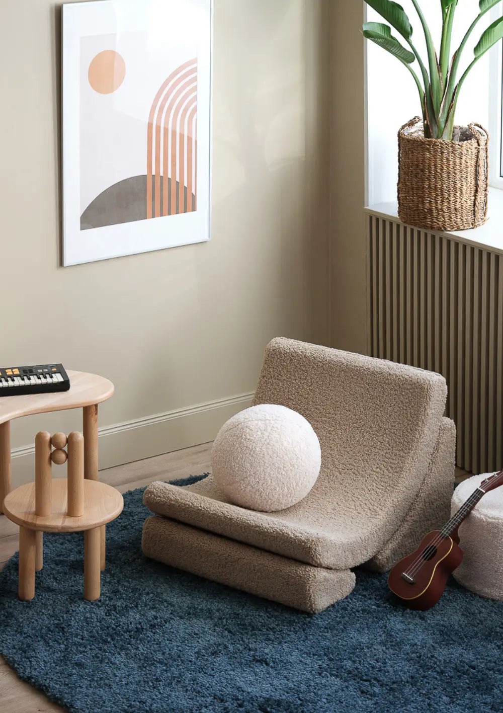 Der beigefarbene Wigiwama Kindersessel Moon Chair Teddy Biscuit steht zusammen mit einem runden Plüschkissen auf einem blauen Teppich in einem stilvoll eingerichteten Kinderzimmer.