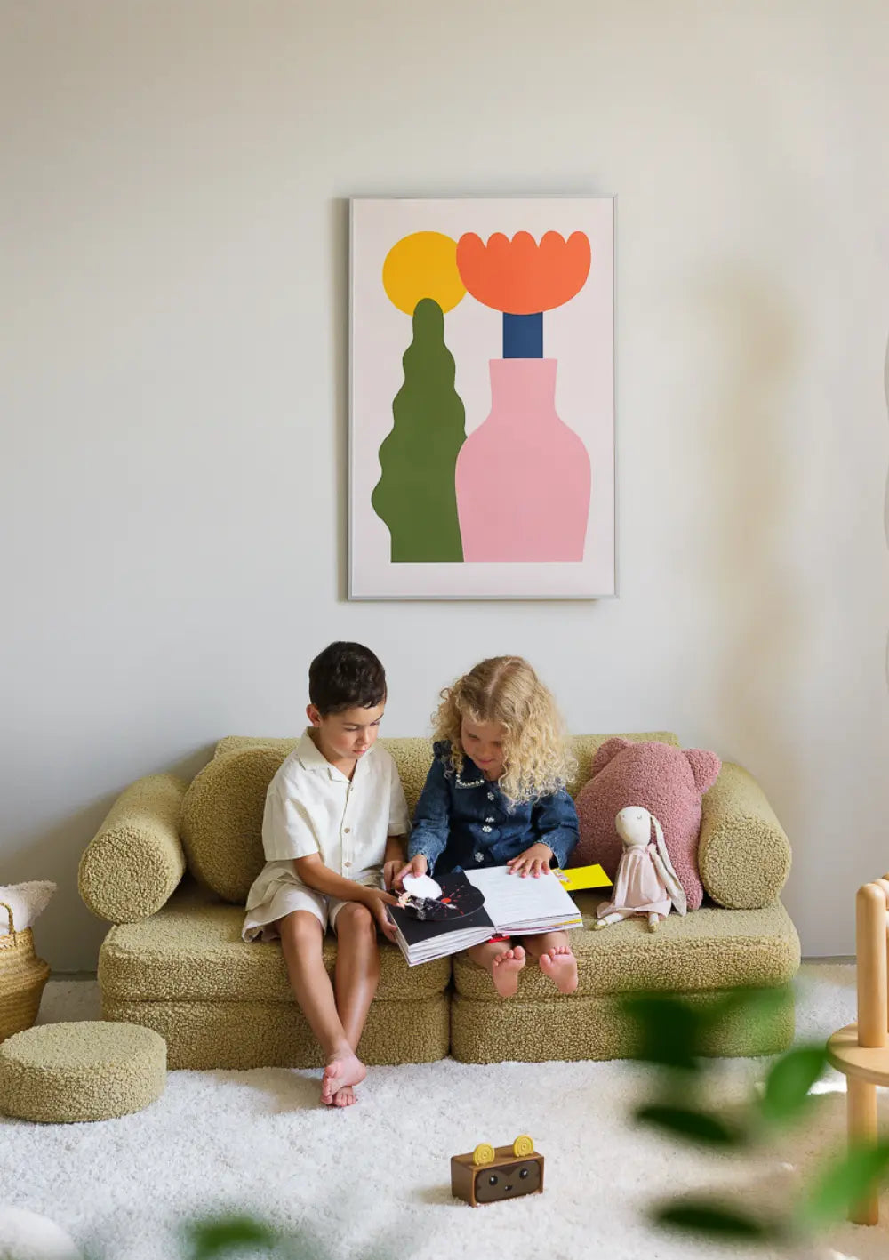 Zwei Kinder sitzen zusammen auf dem olivegrünen modularen Wigiwama Kindersofa Teddy Matcha und blättern in einem Bilderbuch.