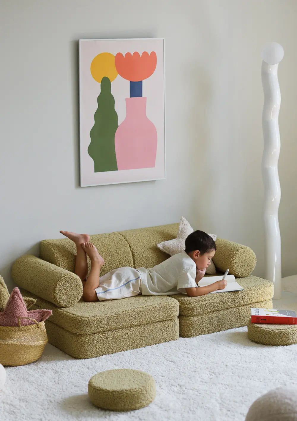 Ein Junge liegt bauchlings auf dem olivegrünen modularen Wigiwama Kindersofa Teddy Matcha aus Plüsch und schreibt in ein Heft.
