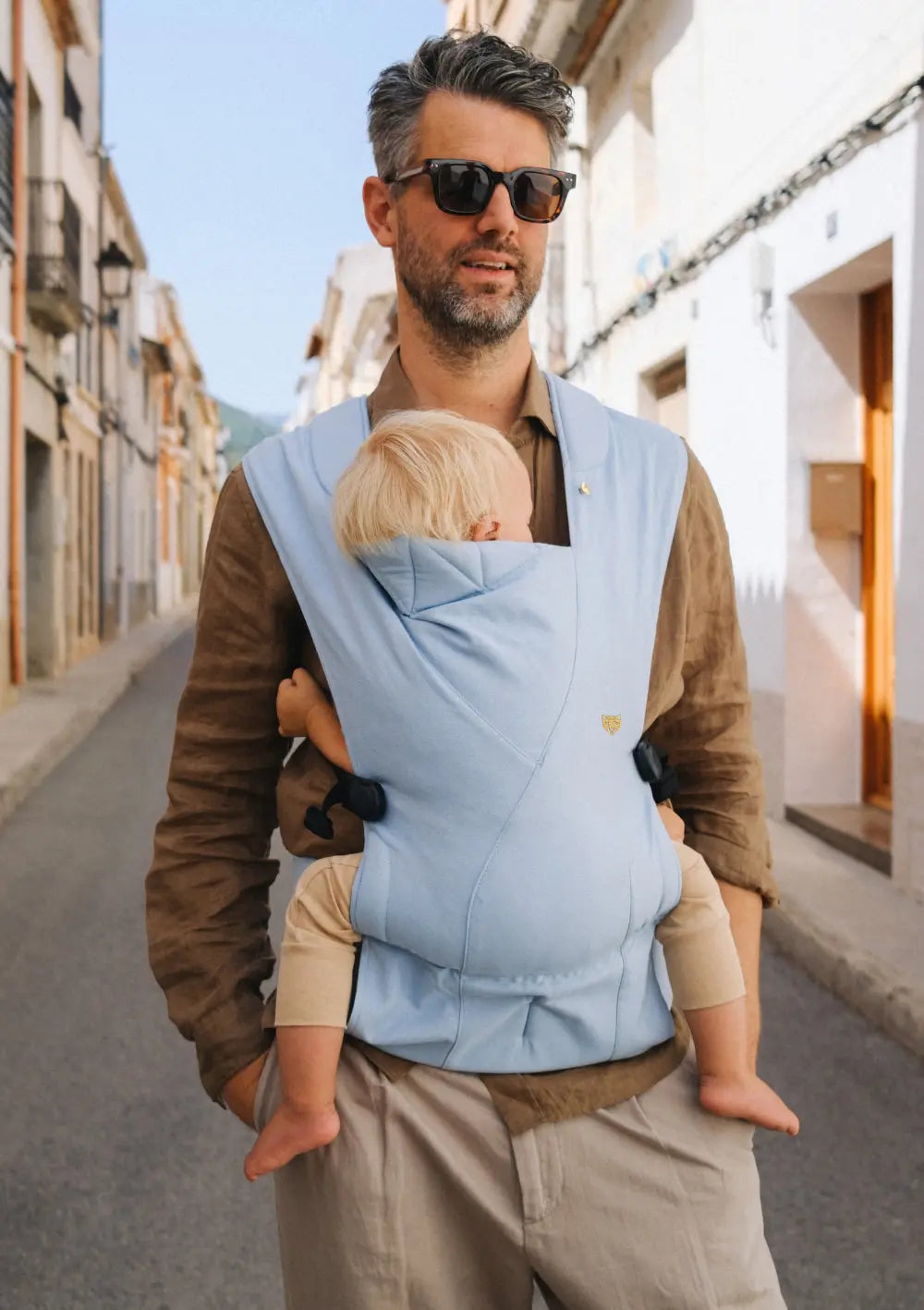 Ein Vater mit Sonnenbrille läuft durch eine Straße und trägt sein Baby vor der Brust in der hellblauen Wildride Babytrage Firstride Bowie Hellblau.